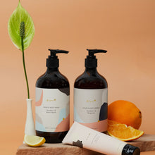 Load image into Gallery viewer, Two bottles of hand and body lotion with a tube of cream on a wooden board against an orange background.