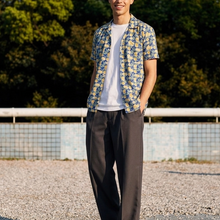 Load image into Gallery viewer, Man wearing a floral shirt and brown pants standing outdoors with trees in the background