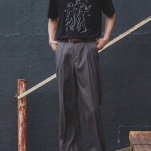 Load image into Gallery viewer, Person wearing a black t-shirt with a graphic design and gray pants against a dark wall.