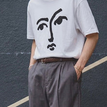 Load image into Gallery viewer, Person wearing a white t-shirt with a face design against a dark textured wall.