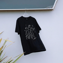 Load image into Gallery viewer, Black t-shirt with white graphic design on a hanger against a white wall.