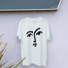 Load image into Gallery viewer, White t-shirt with black face design on a hanger against a light gray wall.
