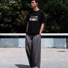 Load image into Gallery viewer, Person wearing a black t-shirt with a graphic design and gray pants standing outdoors.