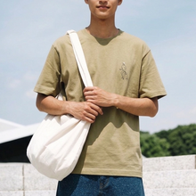 Load image into Gallery viewer, Person wearing a beige t-shirt with a logo, holding a white bag against a clear sky.