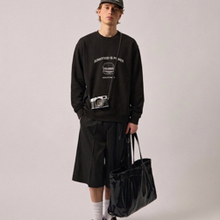 Load image into Gallery viewer, A model wearing a black long-sleeve sweatshirt with a white graphic print, black shorts, white socks, and a black cap, holding a black tote bag against a light gray background.