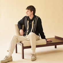 Load image into Gallery viewer, A male model sitting on a chair wearing a black knit polo with contrasting white stripes on the front and back, paired with beige trousers.