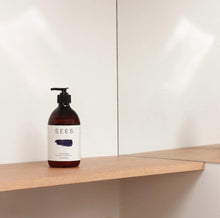 Load image into Gallery viewer, A bottle of SEES Company shampoo with a pump dispenser, featuring a label that indicates an orange and cedar fragrance, positioned on a wooden shelf against a white wall.