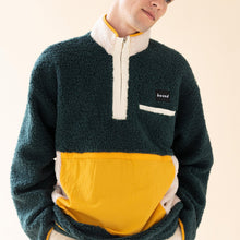 Load image into Gallery viewer, A male model wearing a green and yellow oversized fleece pullover with a ribbed black patch branding above the left breast area. The pullover has a half-zip front and appears to be made of a lightweight material, suitable for winter wear.