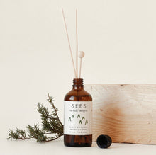 Load image into Gallery viewer, A brown glass scent diffuser bottle with a few drops of liquid, topped with a beige stick, placed on a wooden surface next to green plant leaves. The label has the brand name 'SEES' and the product name 'Mighty Pine'.